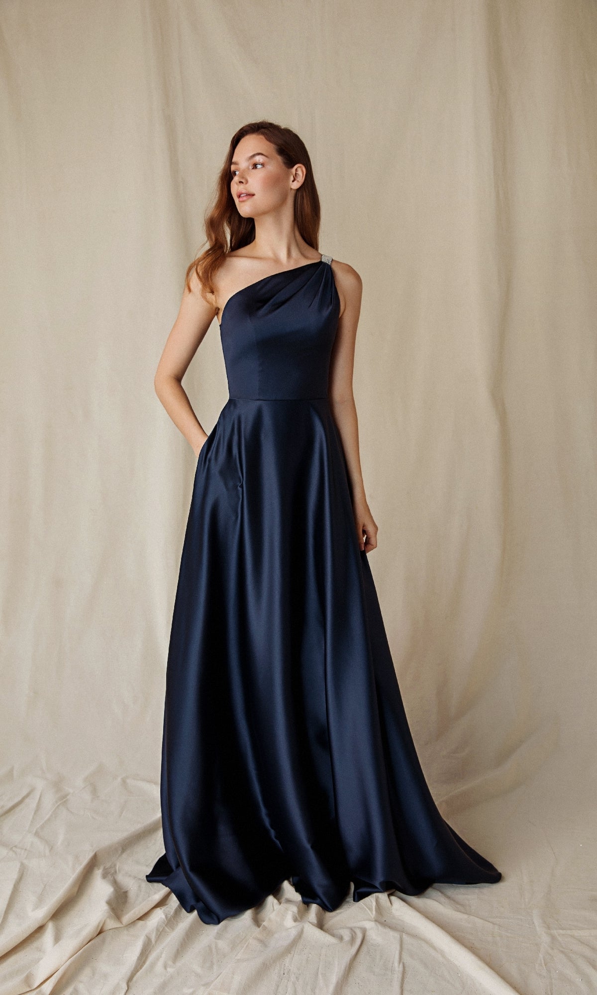 A woman stands in front of a cream-colored fabric backdrop, wearing the Eureka 9025 one-shoulder long satin formal dress in navy blue.